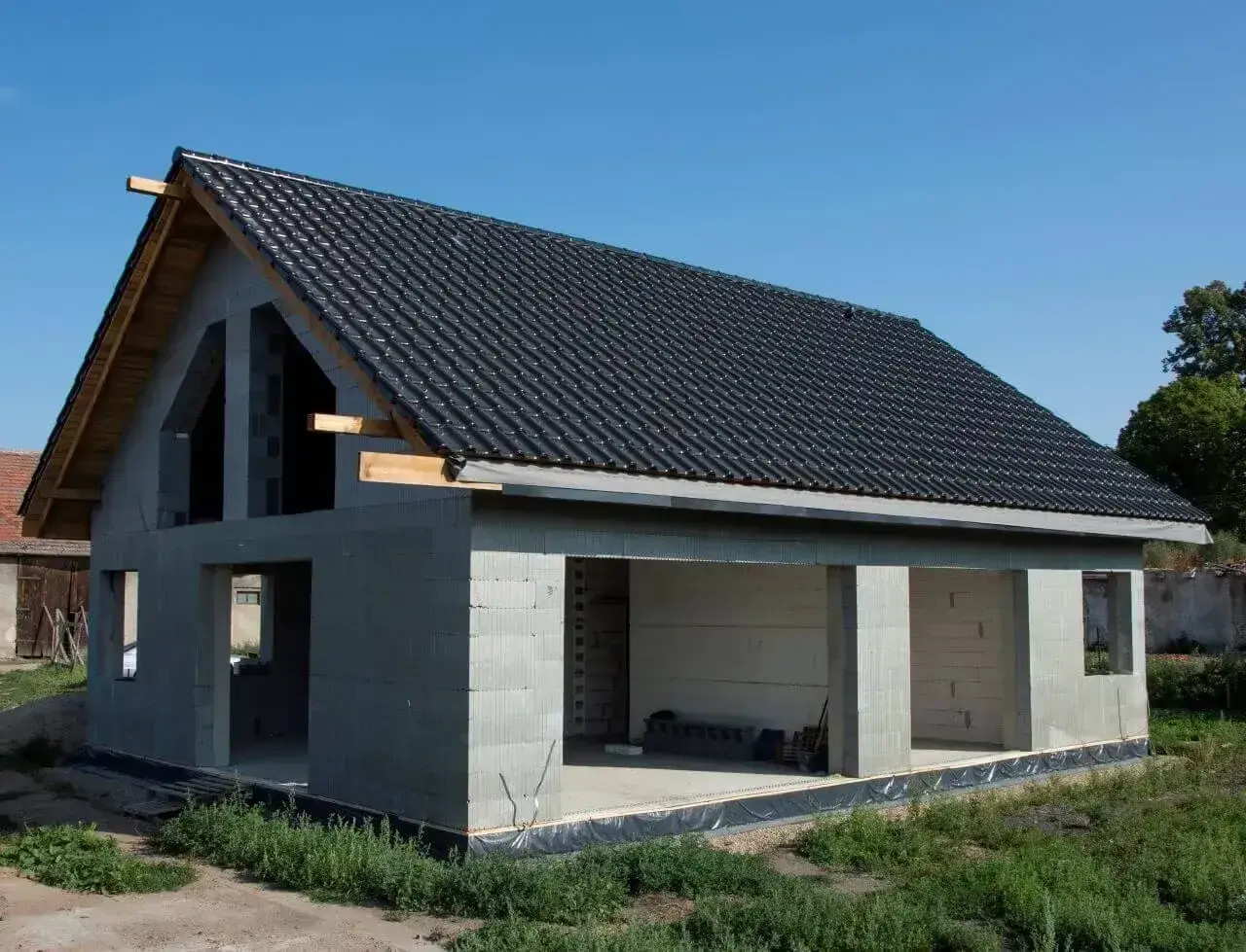 Budynek w stanie surowym otwartym, z betonowych pustaków, z dachem pokrytym czarną dachówką. Widać otwory okienne i drzwiowe.
