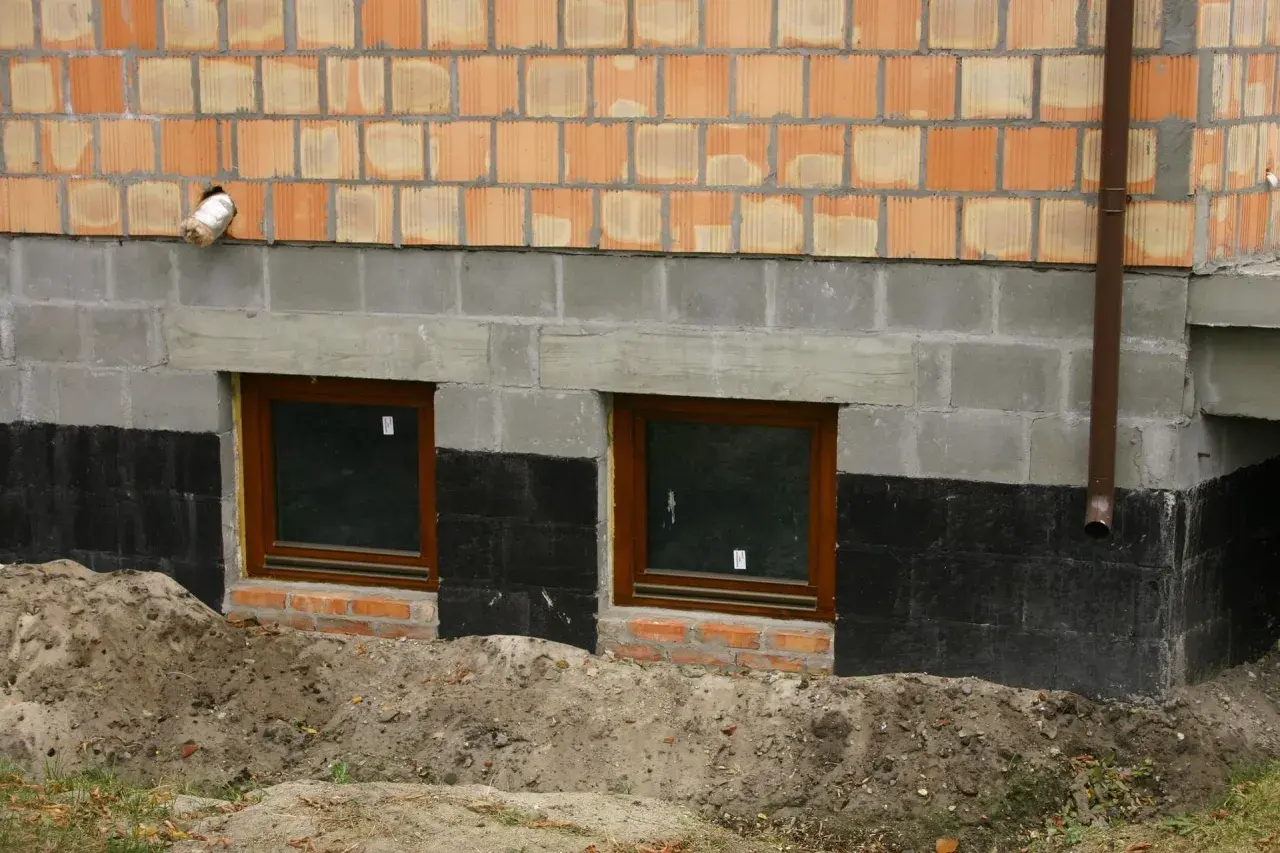 Izolacja fundamentów w starym domu: czarna papa chroni mury przed wilgocią, widać dwa okna piwniczne i ceglaną ścianę.