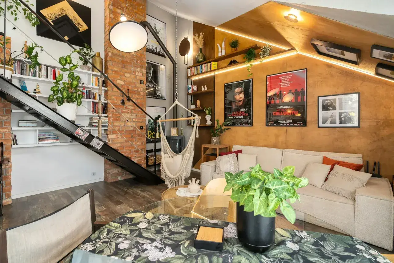 Salon w stylu loftowym z ceglaną ścianą, metalowymi schodami, wiszącym fotelem i wygodną kanapą.