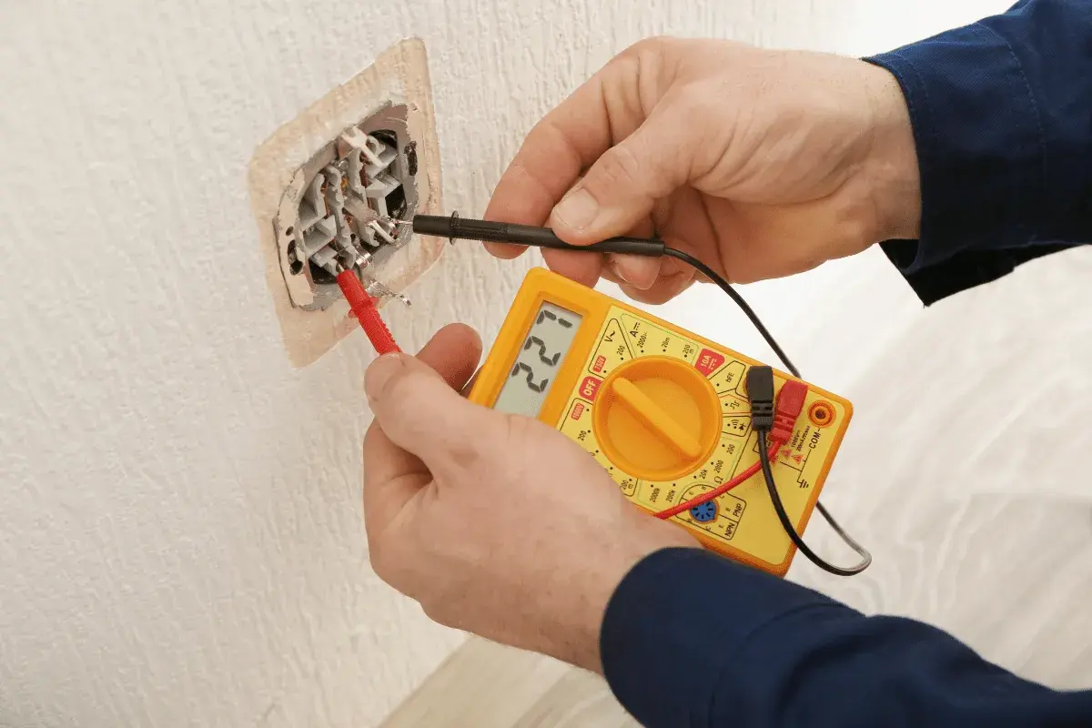 Elektryk sprawdza napięcie w gniazdku za pomocą żółtego multimetru. Wynik pomiaru to 221V.
