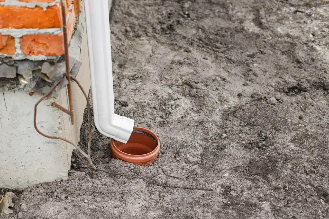 Biała rura spustowa odprowadza wodę z rynien do gruntu, do ceramicznej studzienki.