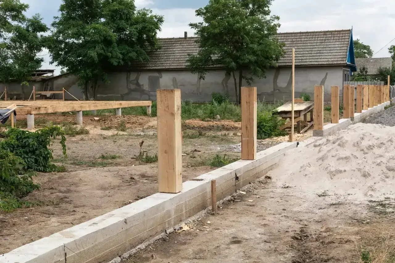 Solidny fundament pod ogrodzenie z drewnianymi słupkami, w tle budowa domu.