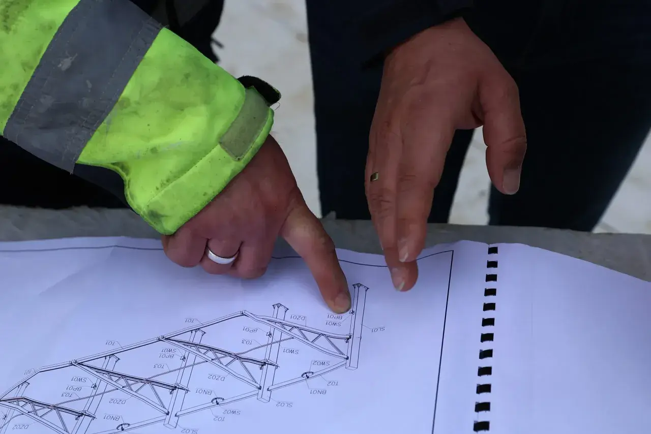 Rysunek techniczny konstrukcji stalowej, omawiany podczas zgłoszenia budowy. Dłonie wskazują na szczegóły projektu.