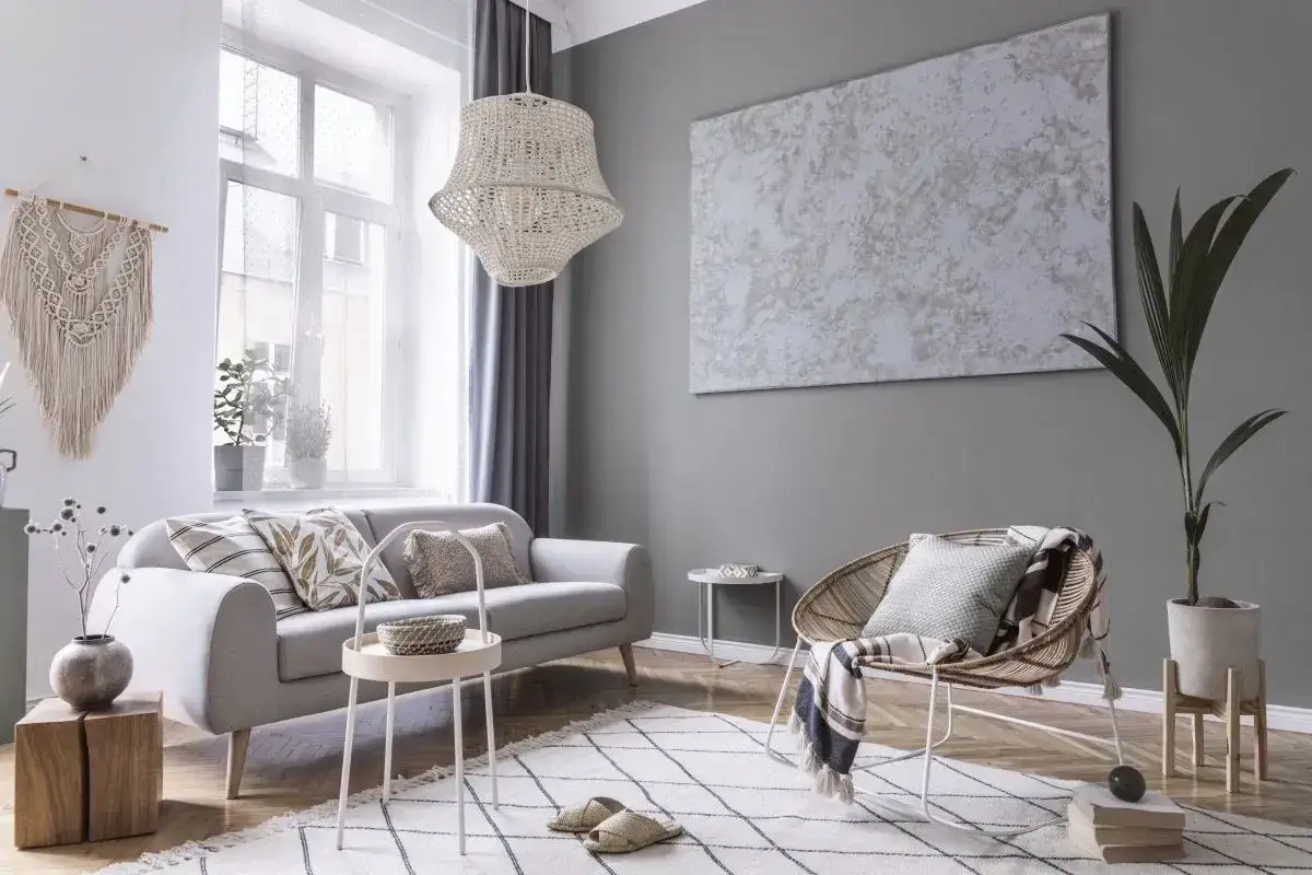 Przytulny salon z szarą kanapą, plecionym fotelem i makramą na ścianie. Dodatki tworzą ciepłą atmosferę.