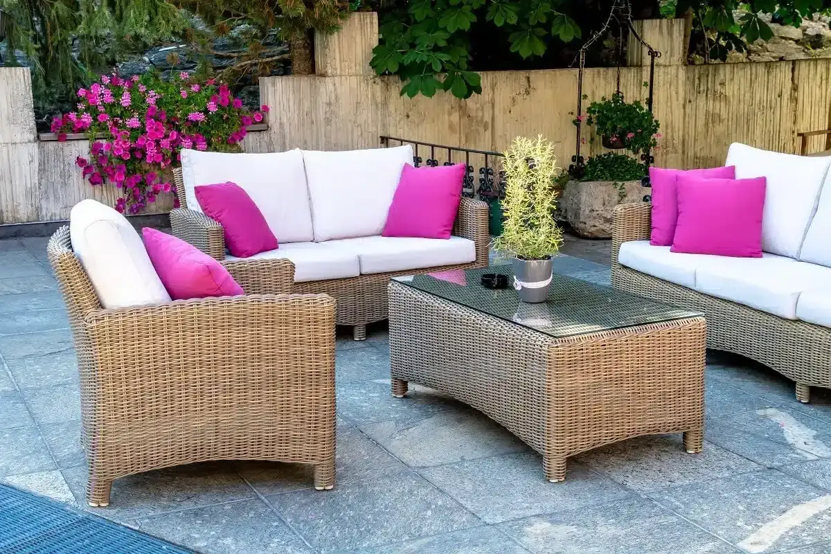 Wygodne, wodoodporne meble ogrodowe z technorattanu: sofa, fotel i stolik kawowy z szklanym blatem, ozdobione różowymi poduszkami.
