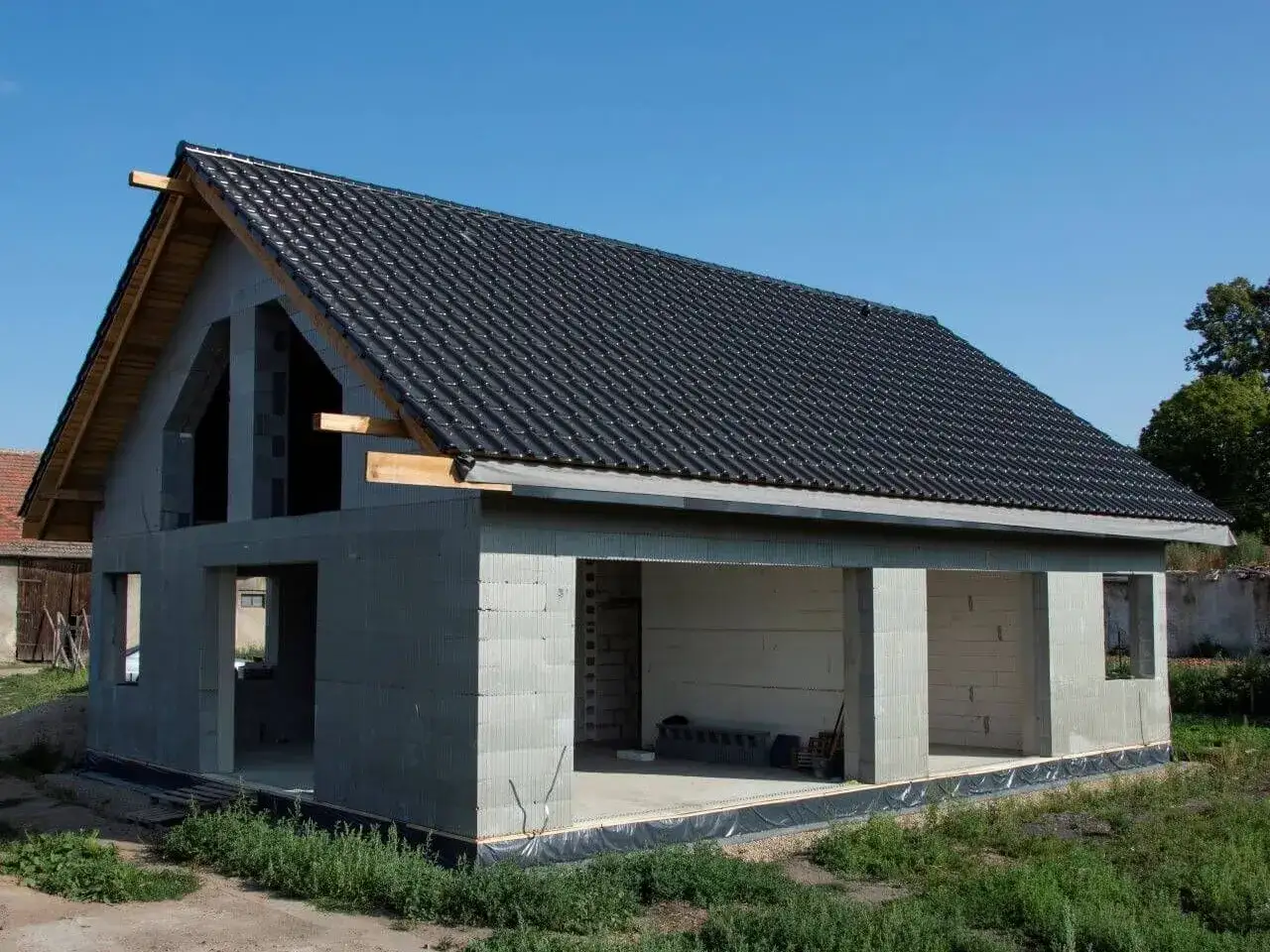Dom w stanie surowym, z betonowych pustaków, z czarnym dachem. Widać otwory okienne i drzwiowe.